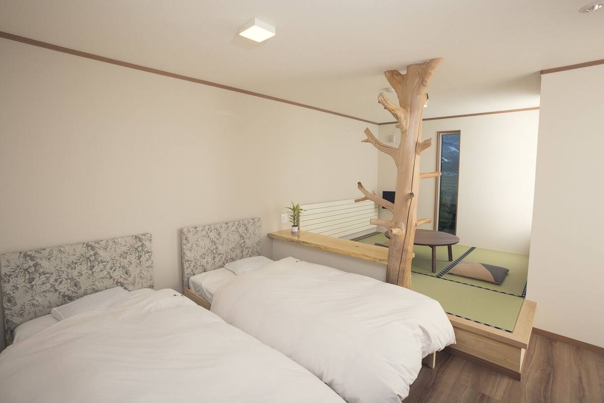 Un dormitorio con dos camas, una escultura de árbol de madera, una mesita con una planta y vistas a una habitación con una silla y una ventana.