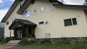 A two-story building with a beige facade, multiple windows, a front porch with steps, and a small garden area. - Ryokan Mokuzin (Hakuba)
