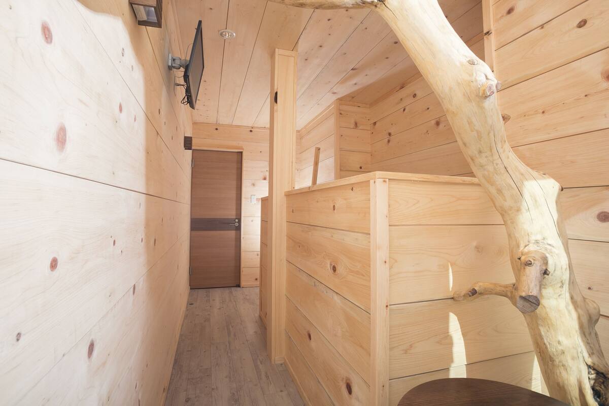 Interior de una cabaña de madera con puerta, banca y estructura de vigas de madera.