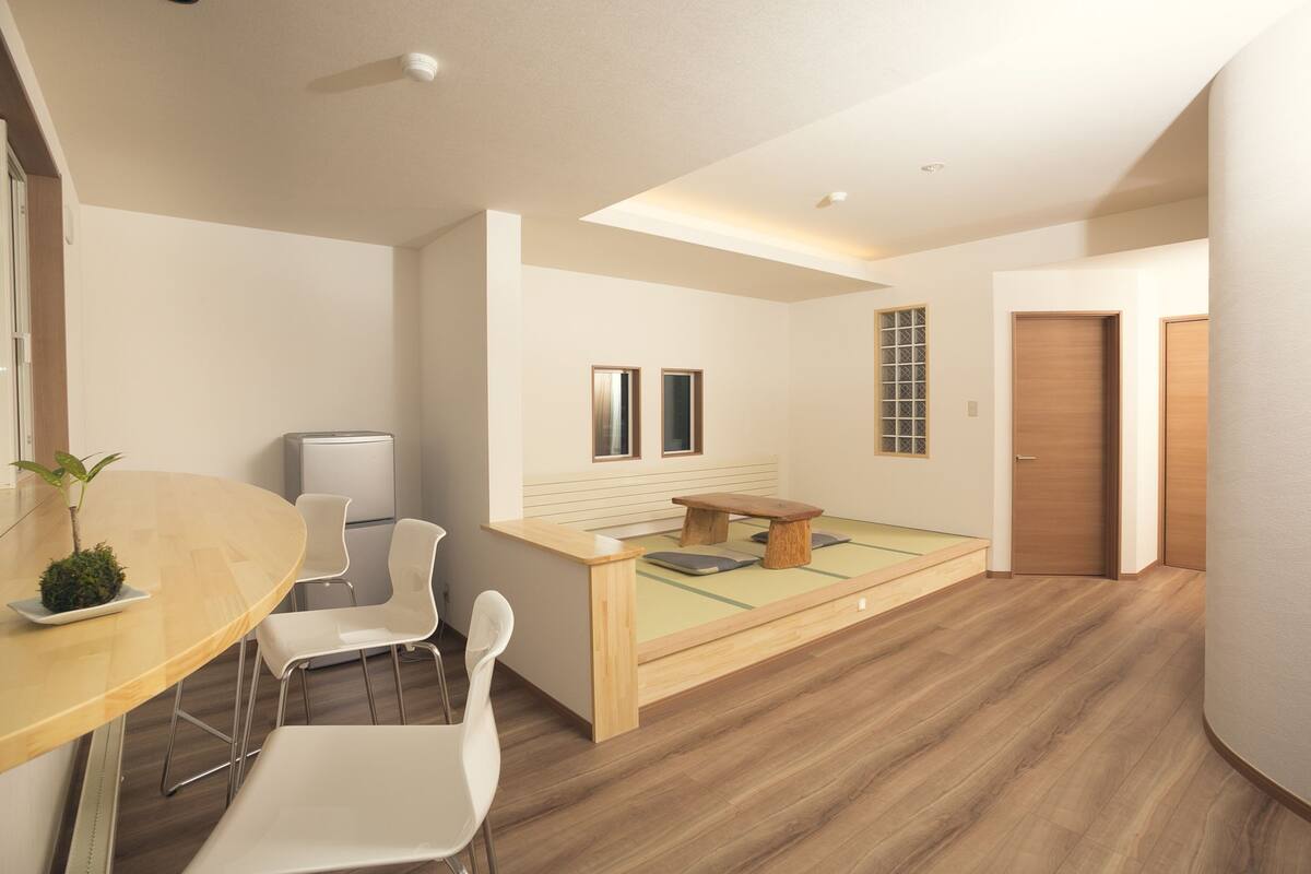 Una habitación moderna y minimalista con una mesa de comedor de madera, sillas blancas, un pequeño banco de madera y una cama con colchón verde.