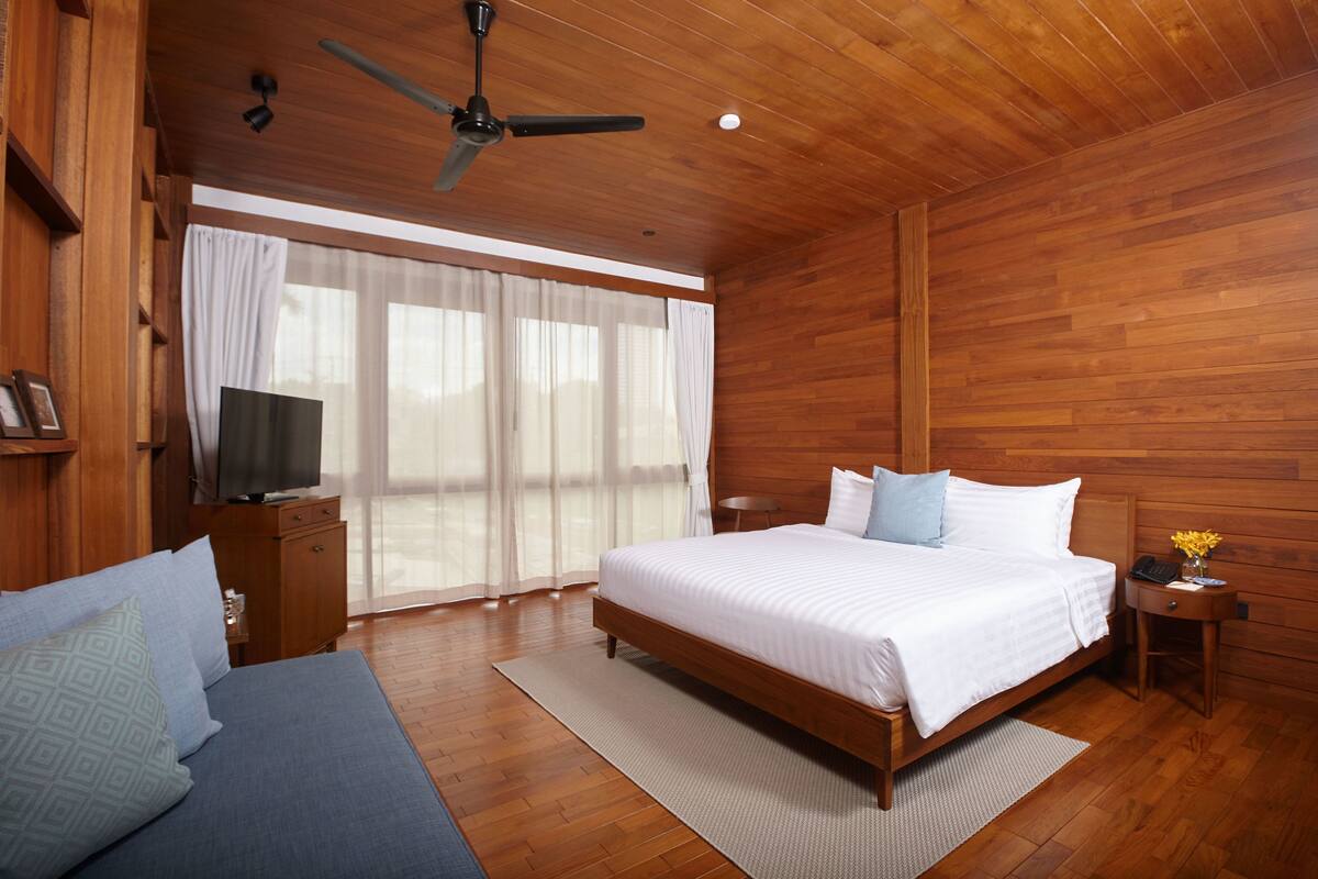 Quarto com painéis de madeira, uma cama, uma televisão sobre um móvel de madeira e uma escrivaninha com um vaso de flores.