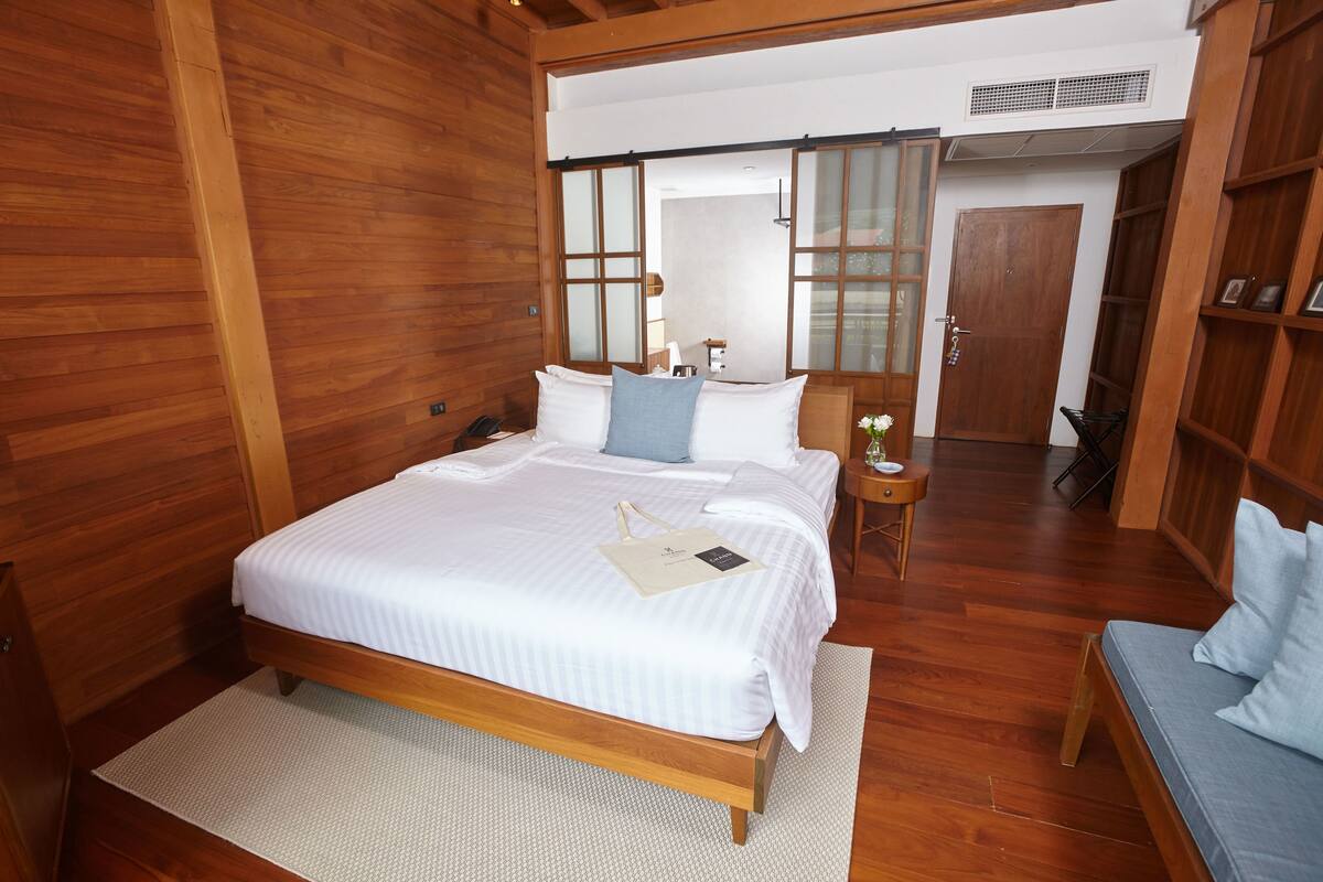 Uma cama com estrutura de madeira, roupa de cama branca e um travesseiro azul, uma mesinha de cabeceira com abajur, e um guarda-roupa de madeira.