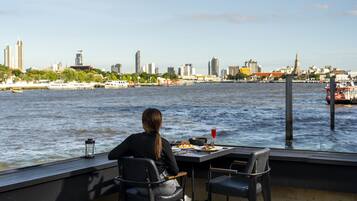 Una mesa de comedor con sillas en una terraza con vista a un río, barcos y el horizonte de una ciudad al fondo.