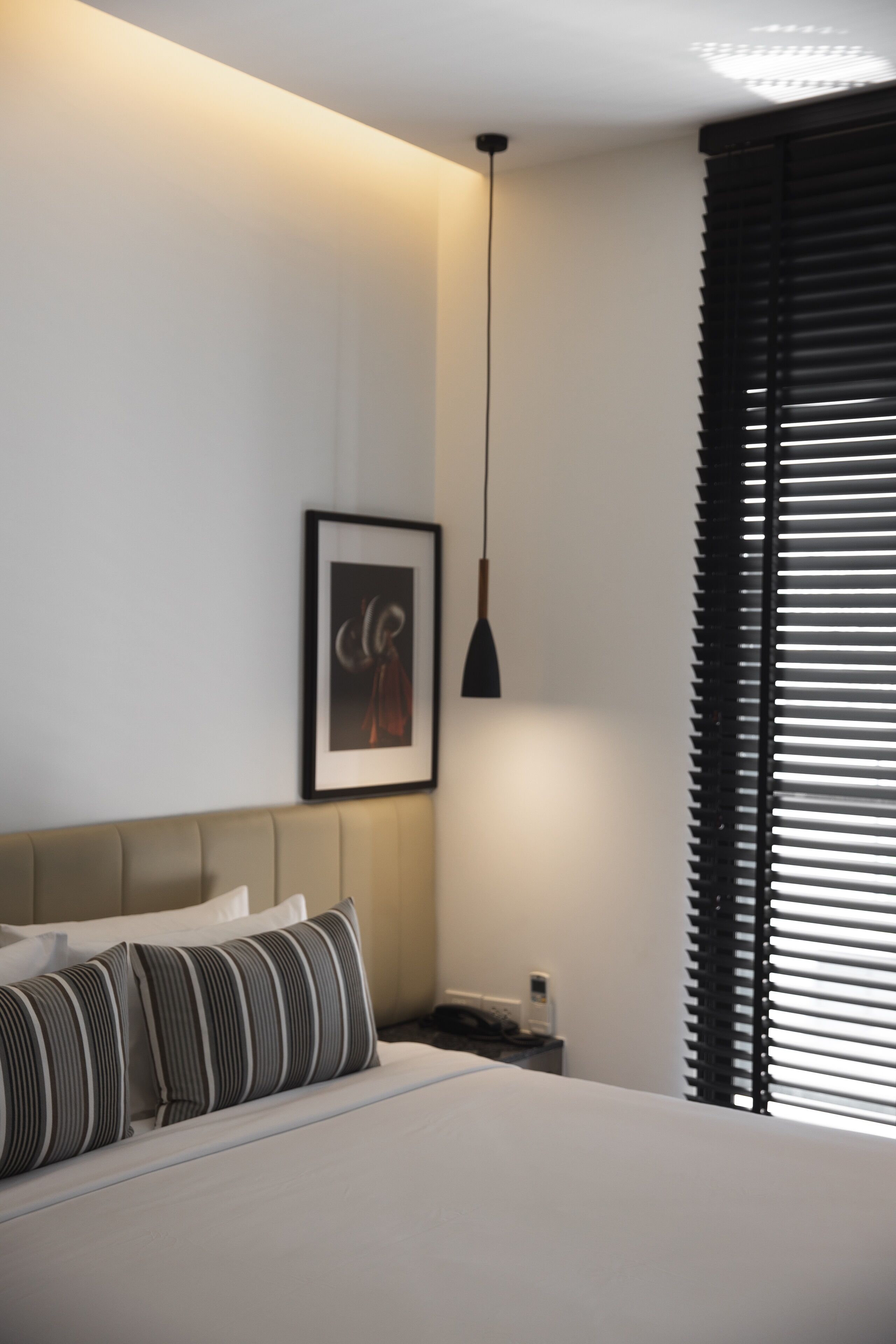 A bedroom with a bed, a framed picture on the wall, a bedside table, and a window with blinds.