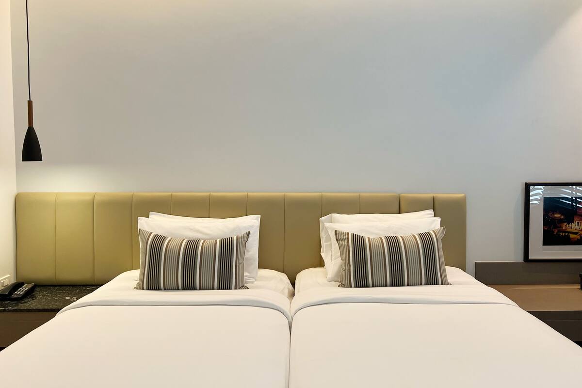 Hotellihuone, jossa on kaksi sänkyä, vaaleanruskea pääty, valkoiset vuodevaatteet, raidalliset tyynyt, musta riippuvalaisin ja seinällä oleva kehystetty kuva.