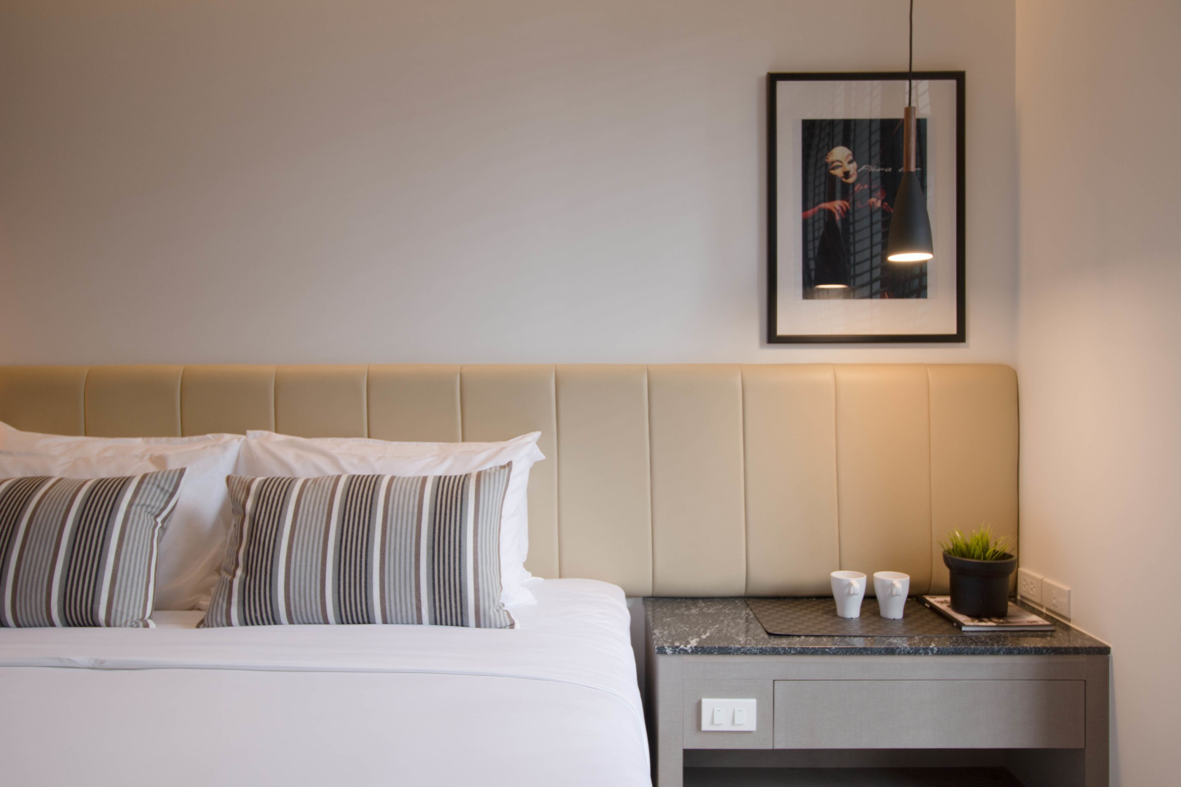 A neatly made bed with striped pillows, a headboard, a bedside table with a potted plant, and a framed picture on the wall.