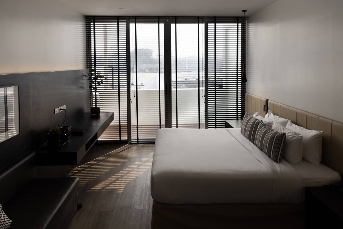 Moderni hotellihuone, jossa on sänky, työpöytä ja suuri ikkuna, jossa on sälekaihtimet.