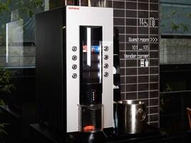 Una cafetera con pantalla digital, una jarra de café y un vaso. Hay carteles que indican números de habitaciones y una estación de café para los huéspedes.