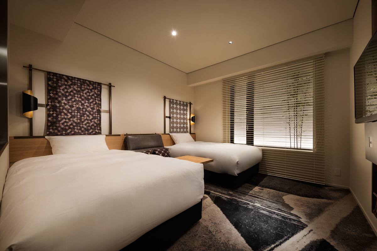 A modern hotel room with two beds, a large window with blinds, a patterned headboard, and a rug.