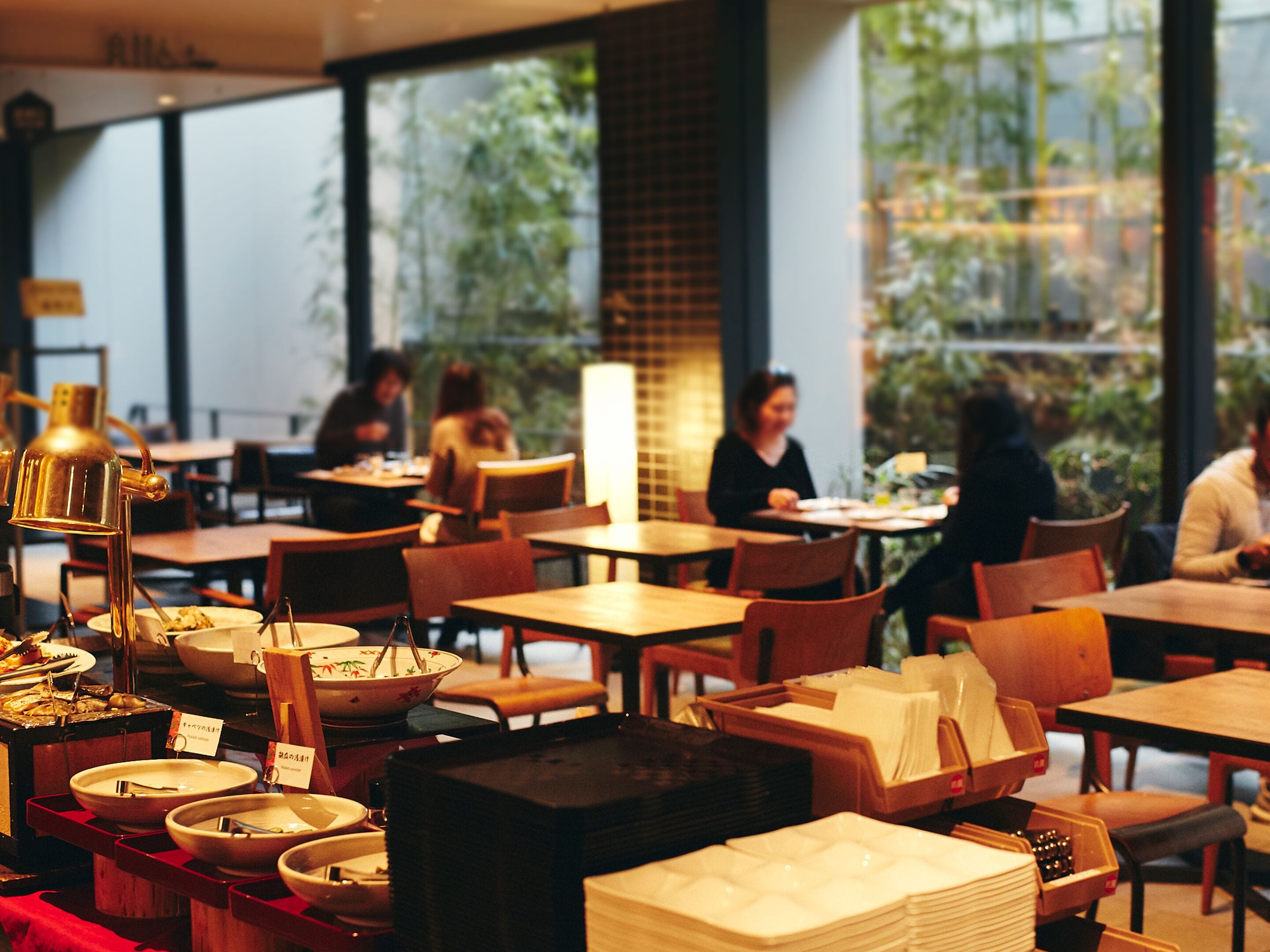 Un restaurant avec des tables et des chaises, un espace buffet avec des plats et des assiettes, et de grandes fenêtres donnant sur un espace verdoyant à l’extérieur.
