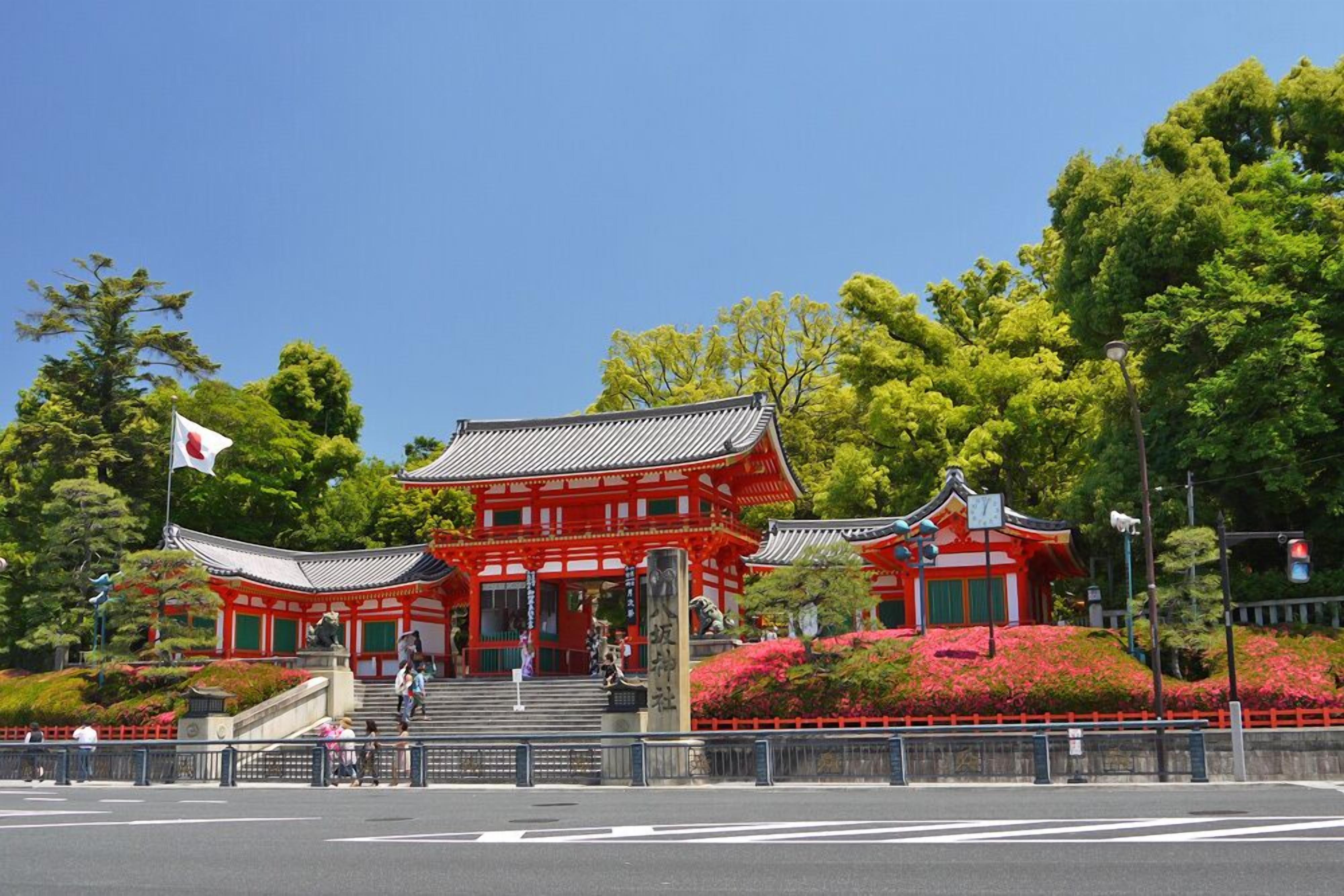 Un bâtiment traditionnel japonais à l’extérieur rouge, entouré d’une végétation luxuriante et surplombé d’un ciel bleu clair.