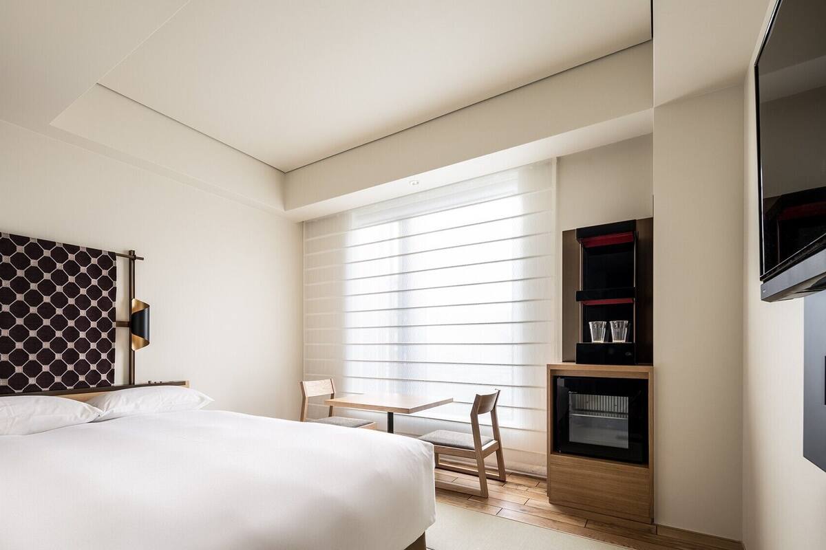 A hotel room with a bed, a desk, a TV, and a window with blinds.