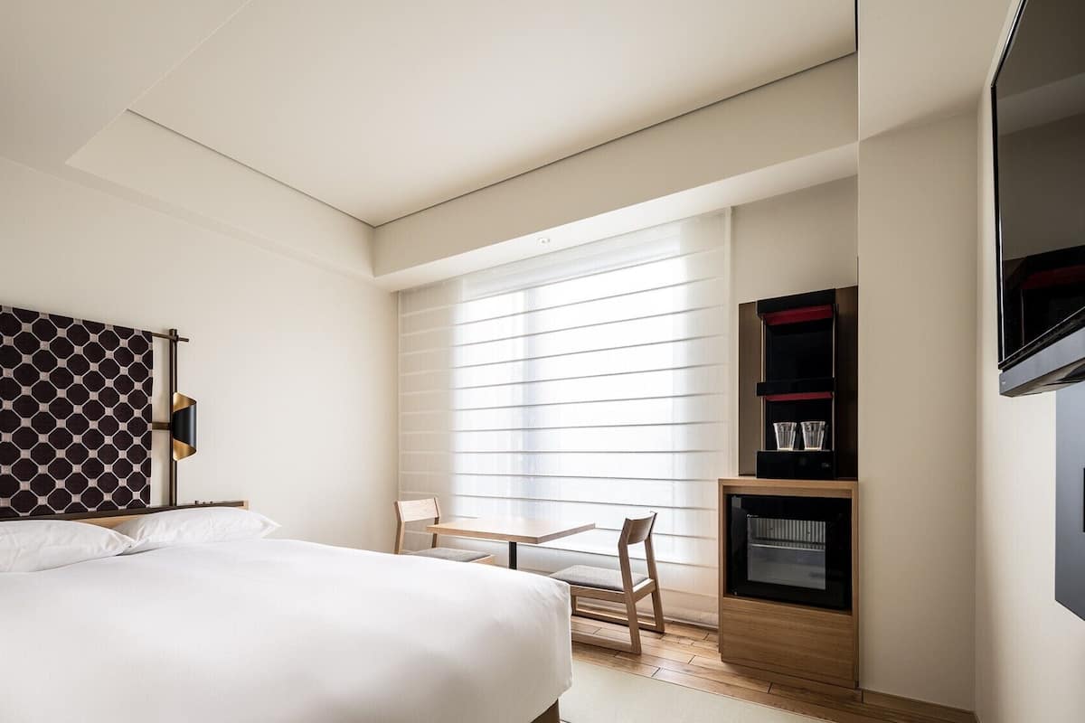 A hotel room with a bed, a desk, a TV, and a window with blinds.