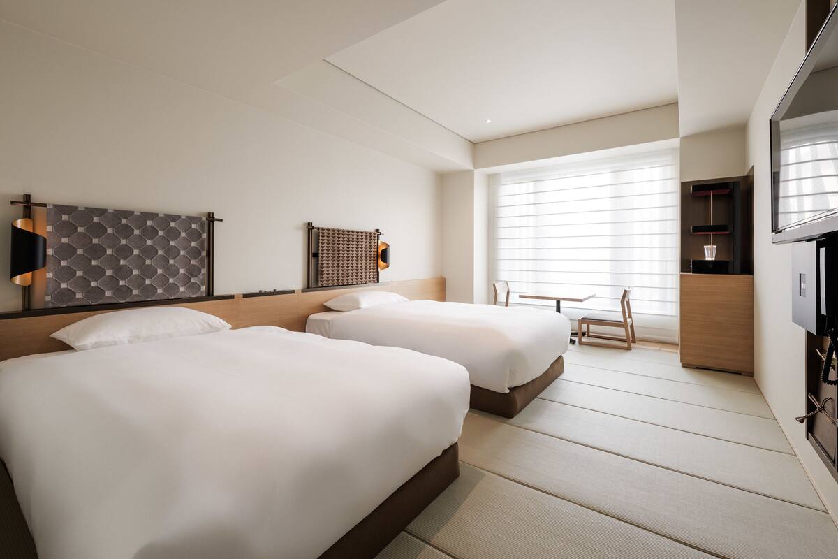 A hotel room with two beds, a TV, a desk, and a window with blinds.
