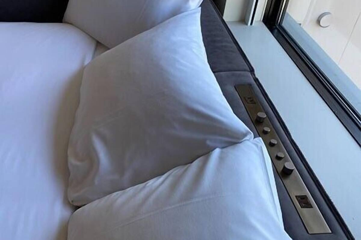 A bed with white bedding and pillows, a window with blinds, and a control panel on the side.