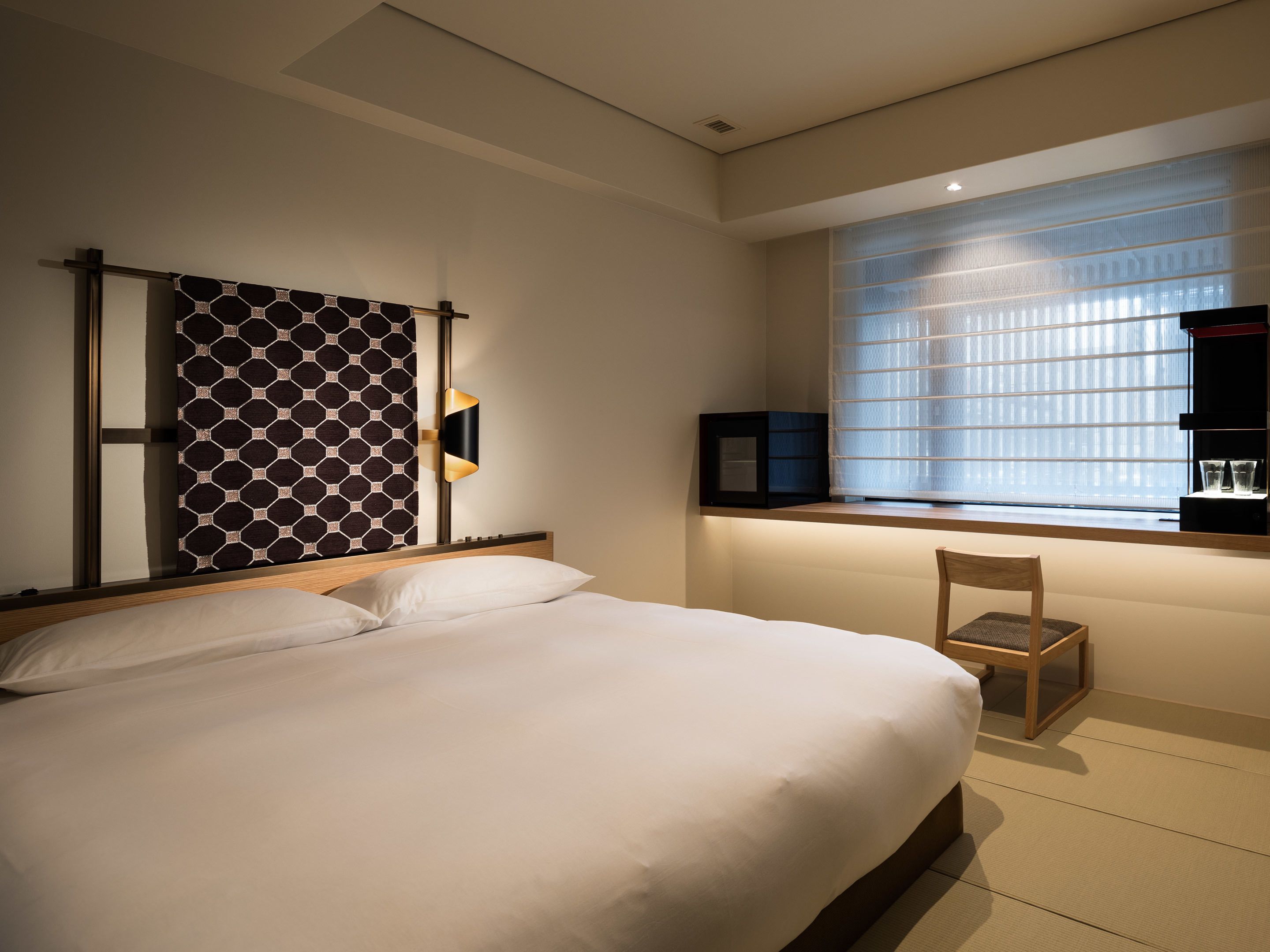 Habitación de hotel con cama, una silla, un estante en la pared con vasos y una ventana con persianas.