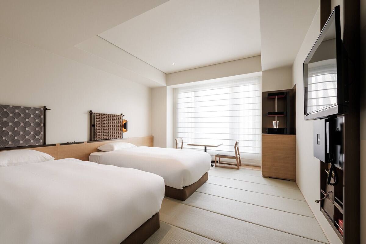 A hotel room with two beds, a TV, a desk, and a window with blinds.
