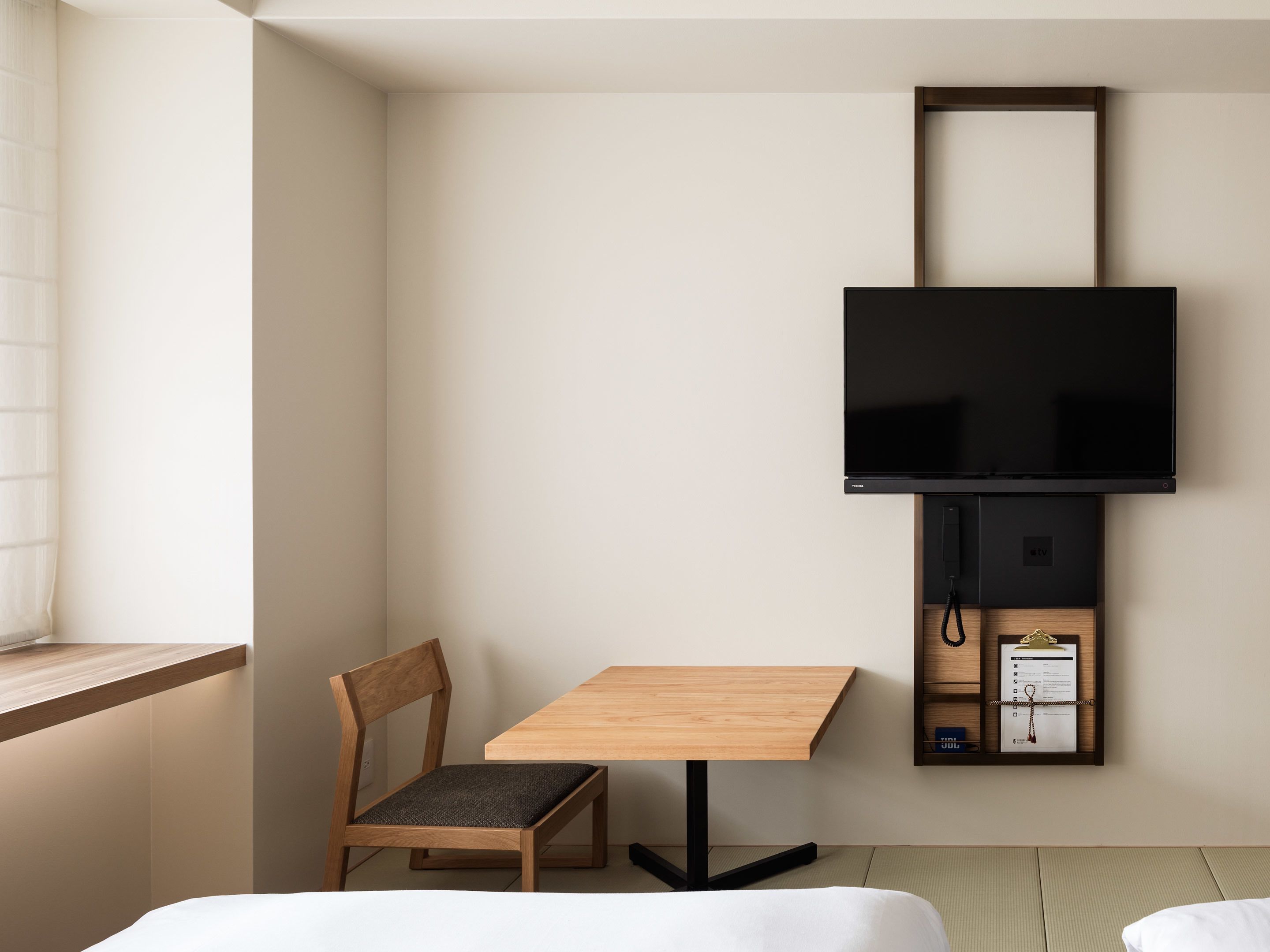 A minimalist room with a wooden table, a single chair, a flat-screen TV mounted on the wall, and a small shelf with a TV remote and a clipboard.