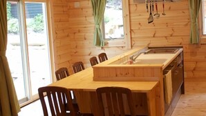 Interior de una cabaña de madera con una amplia mesa de comedor, sillas, una zona de cocina con pileta y utensilios colgantes.