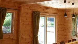 Interior de una cabaña de madera con comedor, ventanas amplias con cortinas e iluminación colgante.