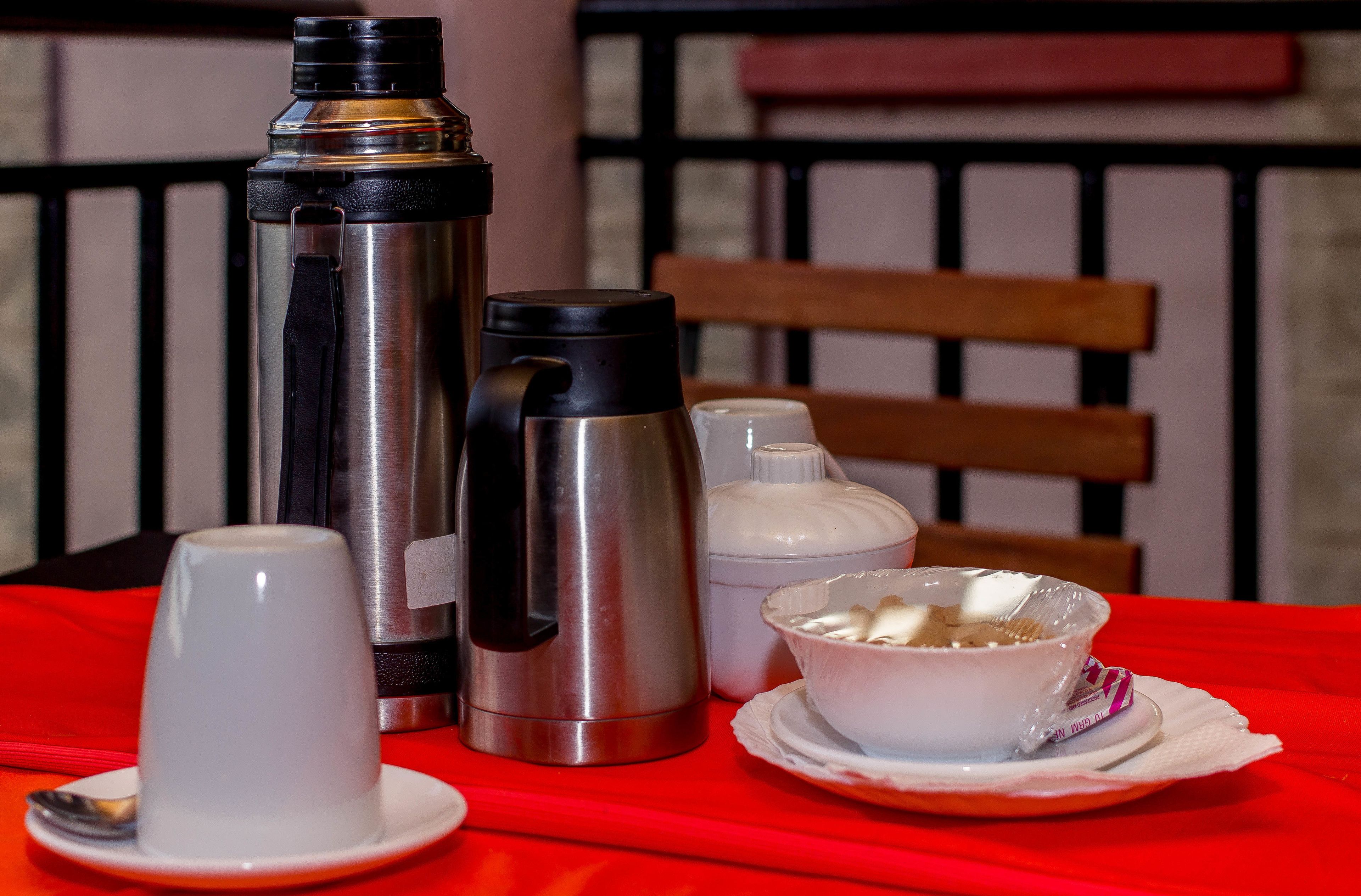 A table with a red tablecloth, a stainless steel thermos, a black thermos, a white mug, a white bowl with food, and a spoon.