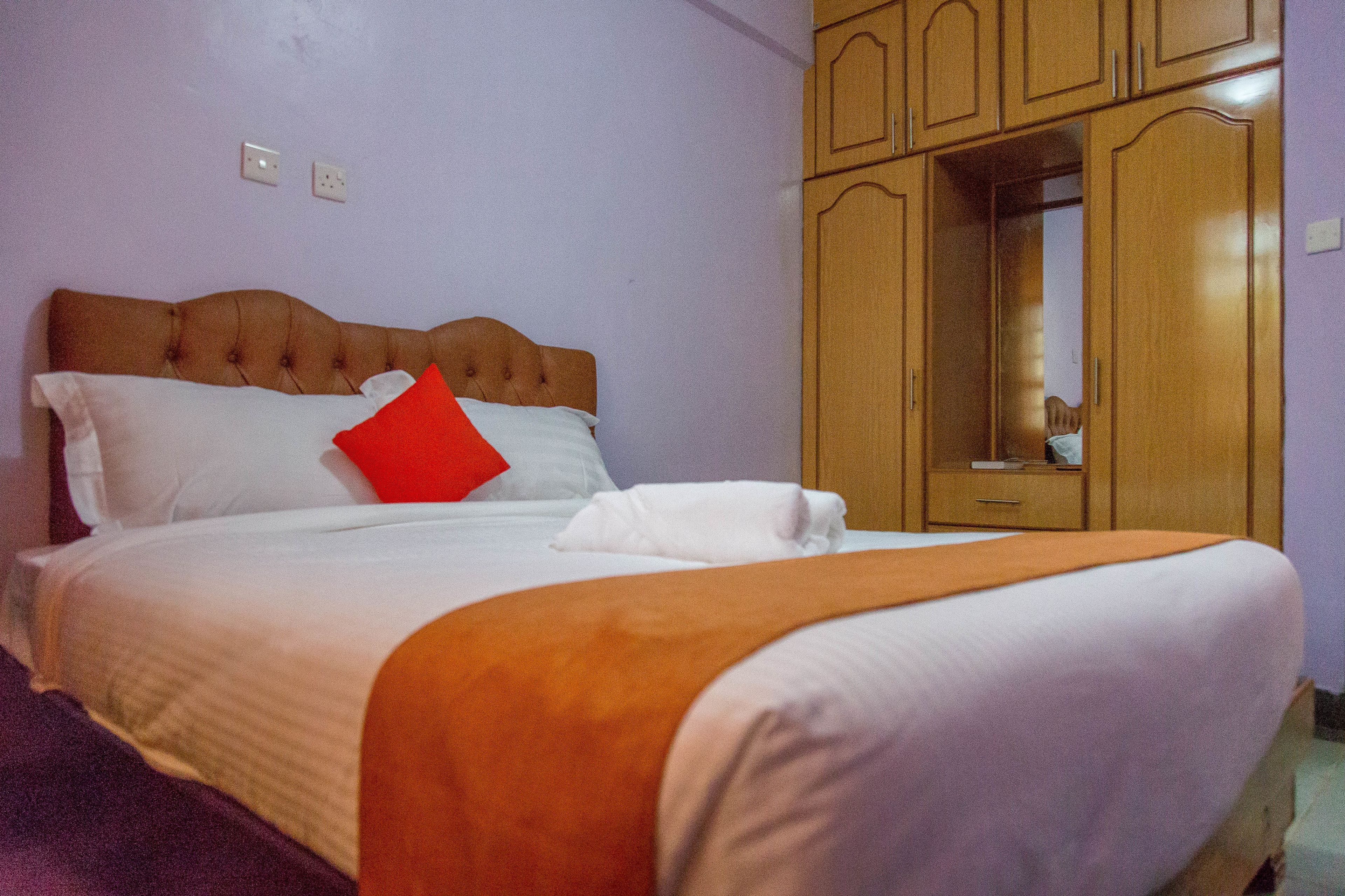 A hotel room with a bed, a red pillow, a white pillow, a white blanket, and a wooden wardrobe.