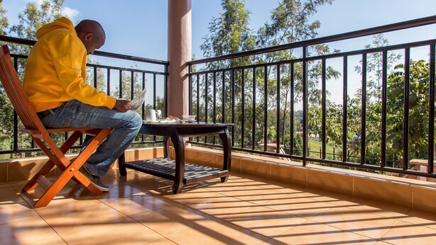 A person seated on a wooden chair on a balcony with a tiled floor, a small table with a cup and saucer, and a railing with a view of trees and hills. - Kuniville Guest House (Nyeri)