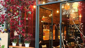 Un restaurant avec une porte vitrée ornée de décorations annonçant le « Bonne et heureuse année du Nouvel An chinois ». À l’intérieur, on trouve des tables et des chaises en bois, des plantes en pot et un mur en briques.