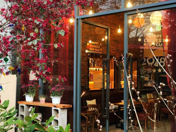 Un restaurante con puerta de cristal que exhibe decoraciones de 'Happy Chinese New Year'. En su interior, hay mesas y sillas de madera, plantas en macetas y una pared de ladrillo.