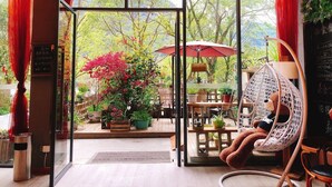 Un espace extérieur accueillant avec des sièges, comprenant une chaise en osier et un parasol rouge, entouré de plantes en pot et offrant une vue sur les arbres.