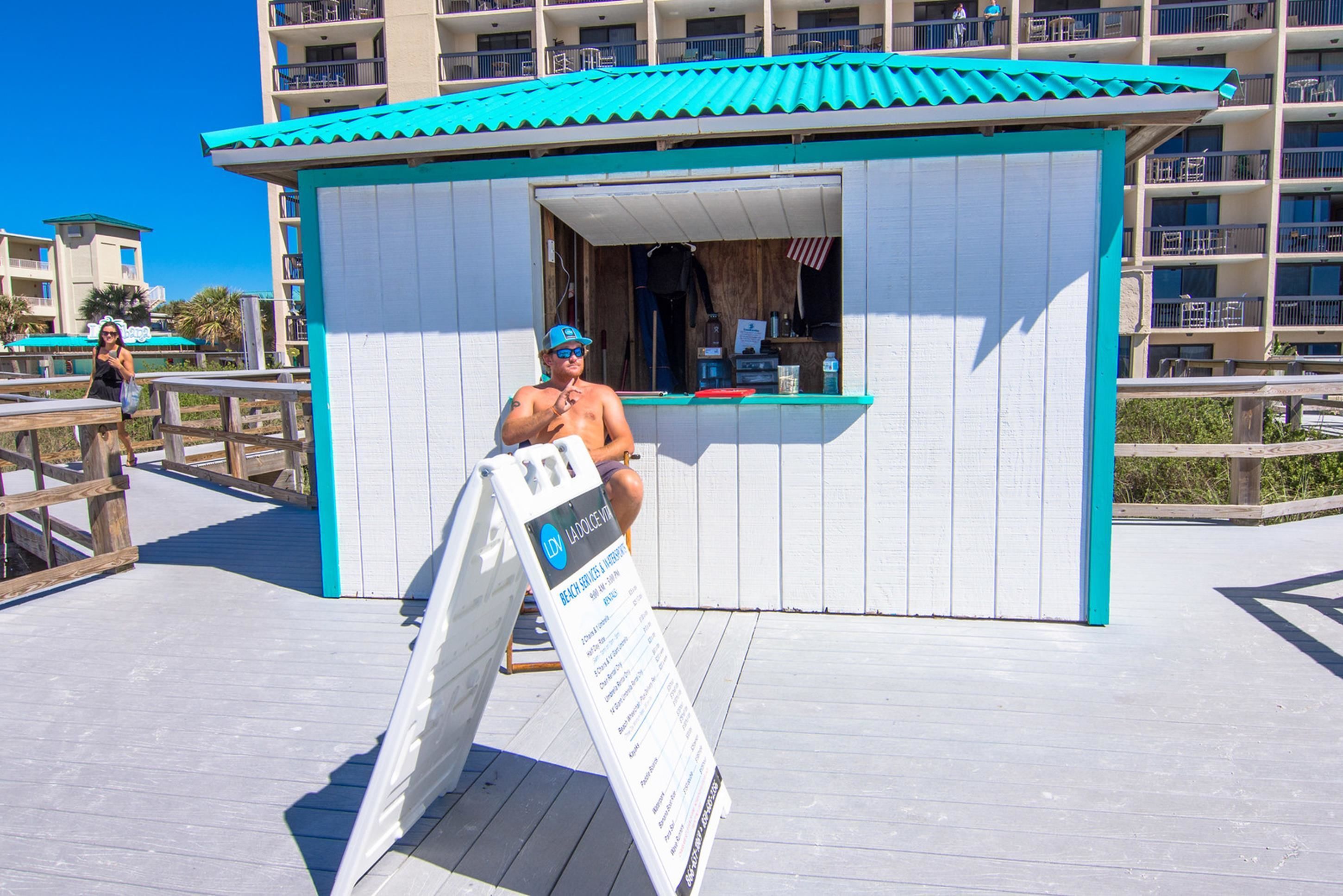 A beachside kiosk with a signboard, wooden deck, and a building with a blue roof in the background.
