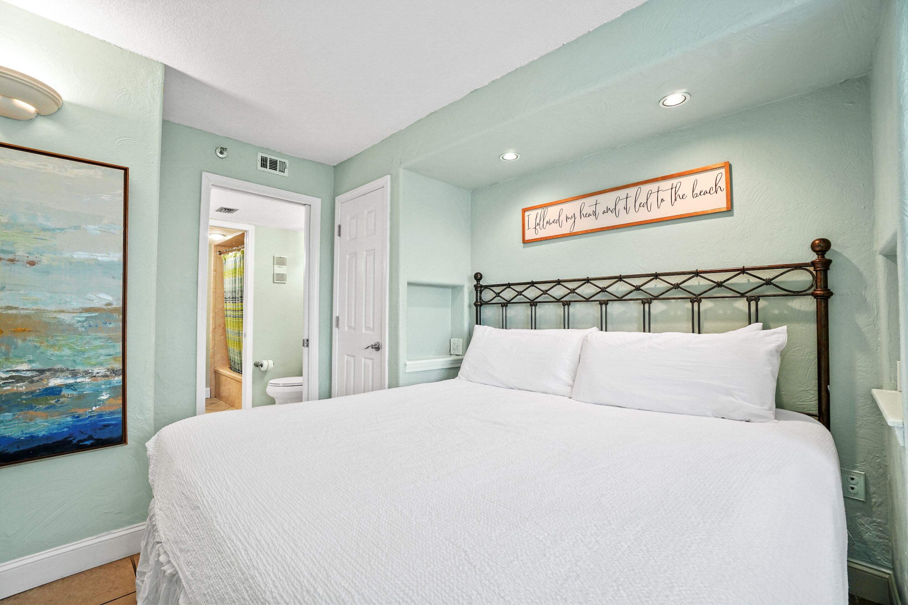 A bedroom with a bed, a painting, a door, and a wall sign.