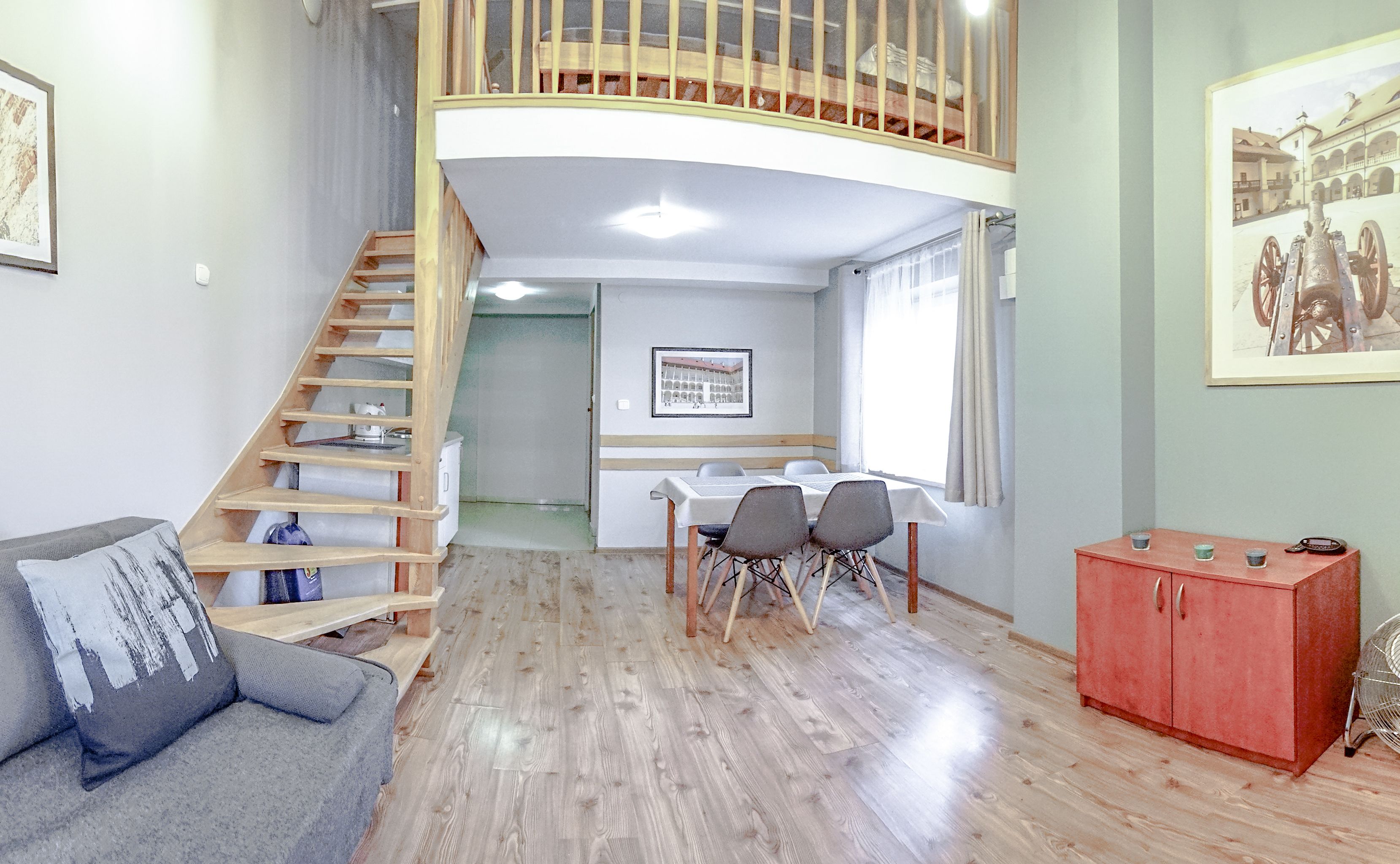 A spacious living area with a wooden staircase, a dining table with chairs, a sofa, a red cabinet, and a framed picture on the wall.