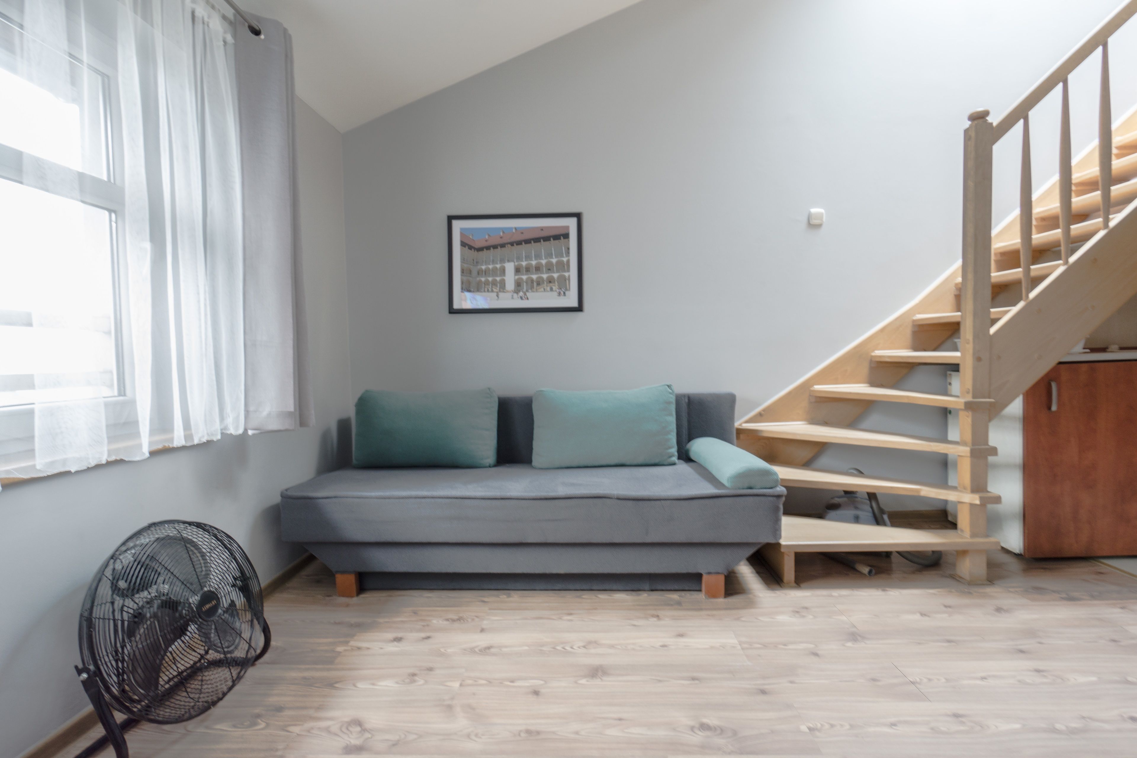 Una habitación con un sofá gris, una escalera de madera, un ventilador y un cuadro en la pared.
