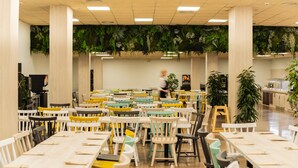A spacious indoor area with multiple tables and chairs, a person walking in the background, and green plants hanging from the ceiling. - Ona Palamós (Palamós)