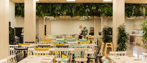 A spacious indoor area with multiple tables and chairs, a person walking in the background, and green plants hanging from the ceiling.
