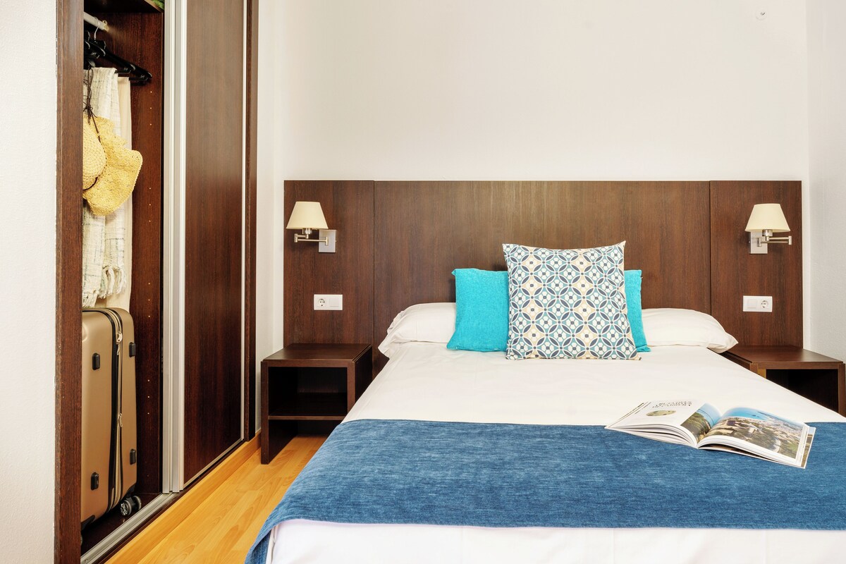 A hotel room with a wooden headboard, two bedside tables, a blue blanket, and a book on the bed.
