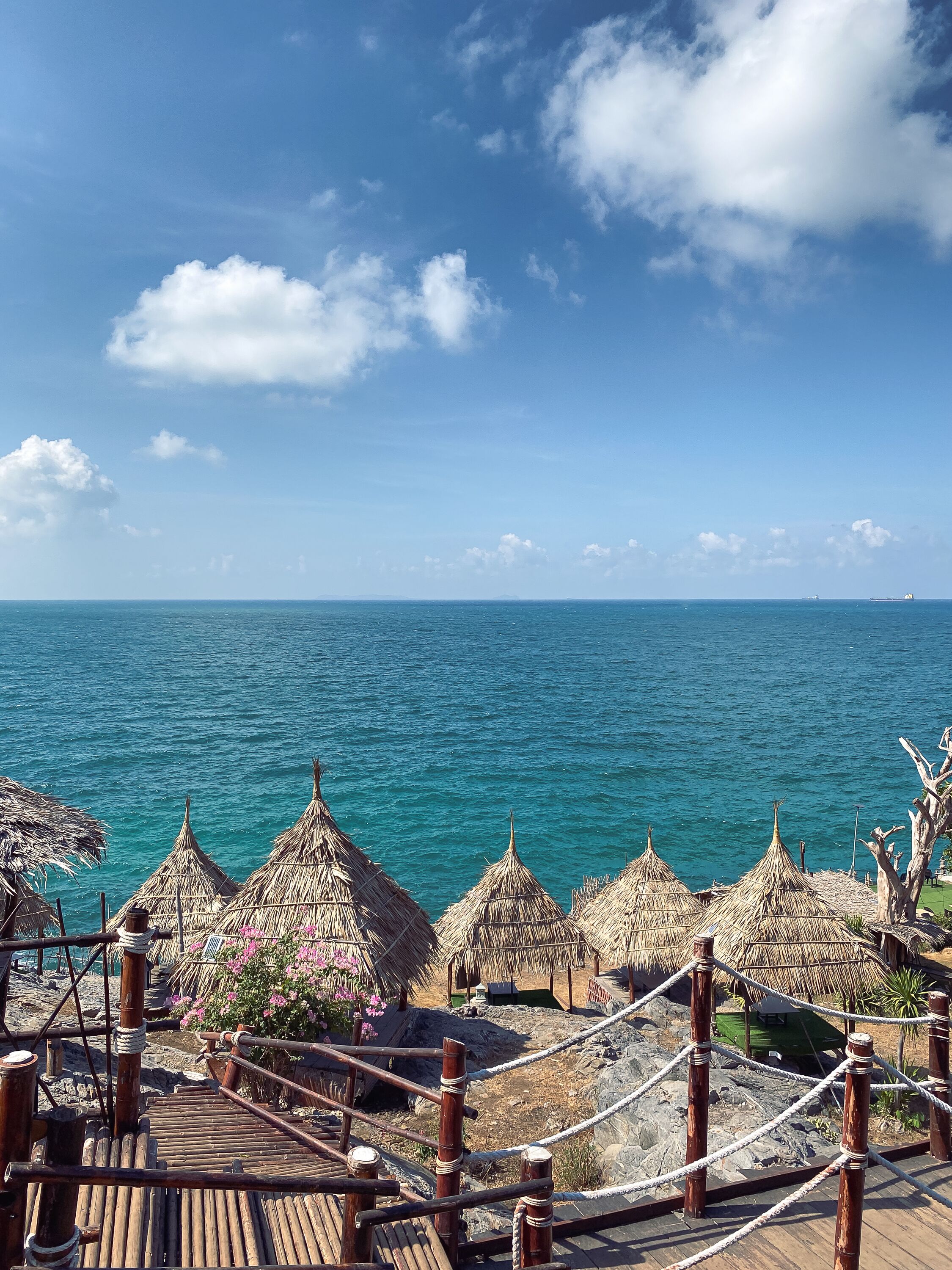 A wooden deck with rope railings overlooking a turquoise sea, with thatched-roof structures and a clear sky.