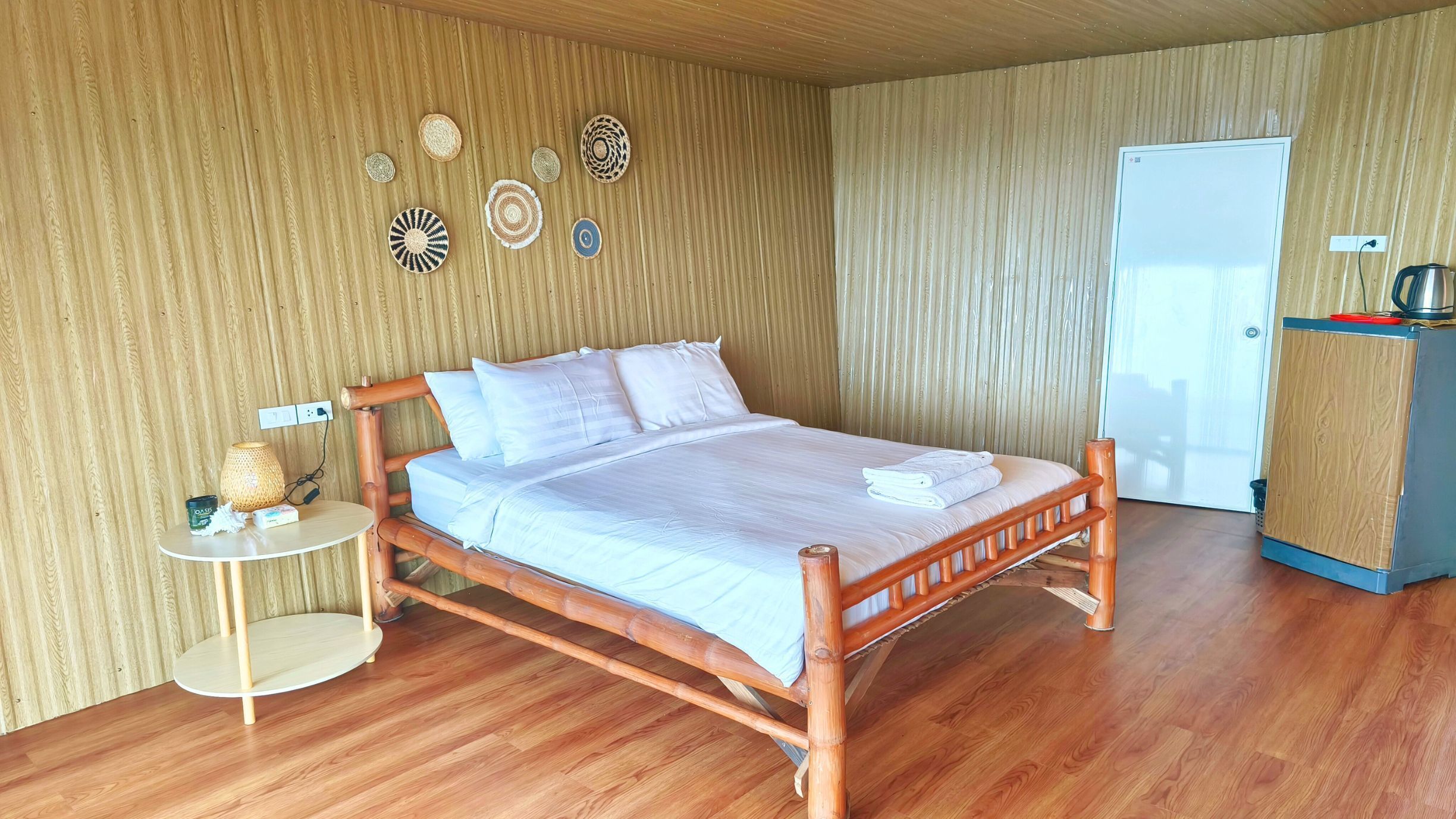 A wooden bed with a white mattress and pillows, a small round table with a lamp, and a wooden cabinet with a kettle on top.