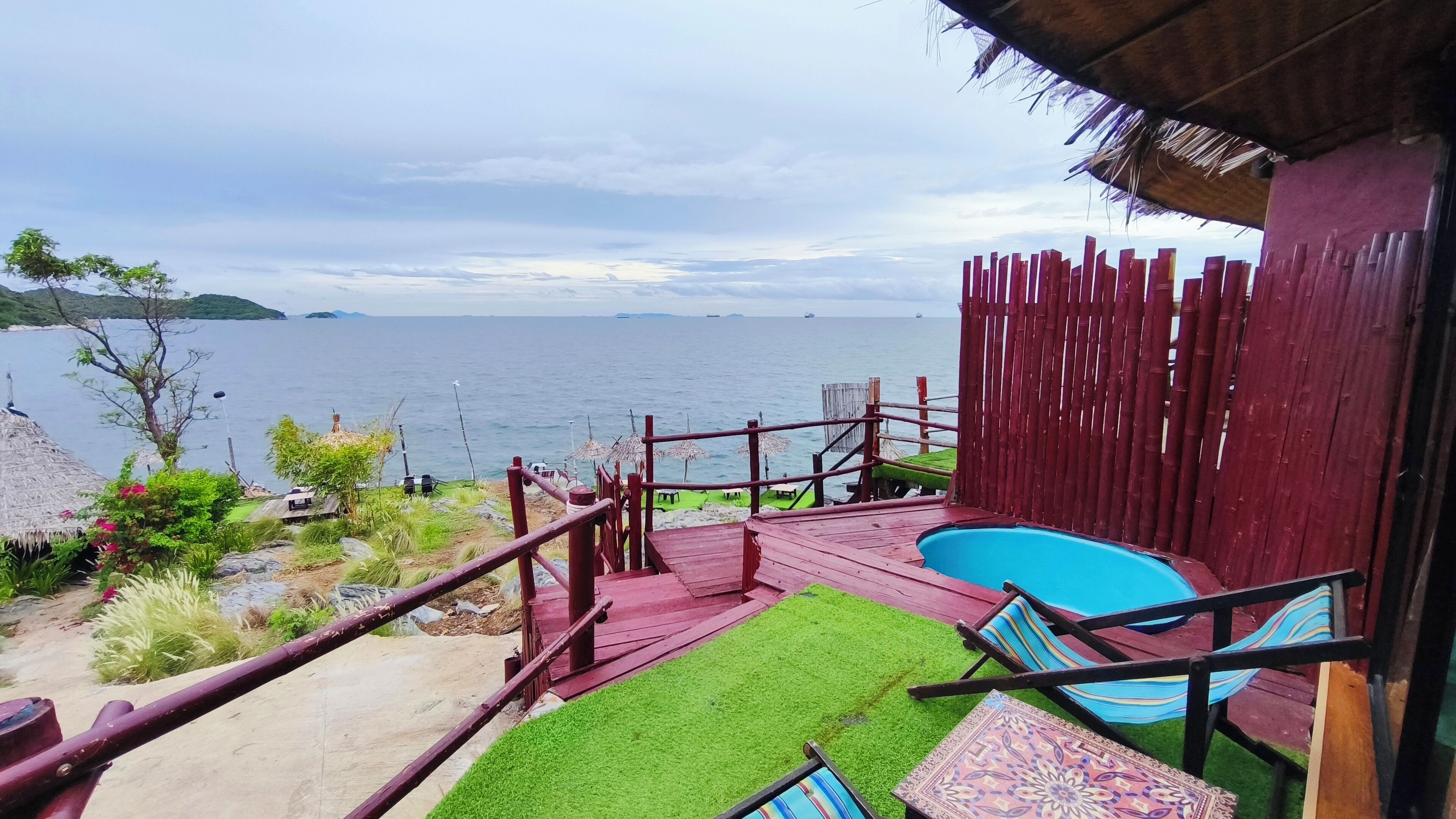 Zona de piscina con vista al mar, terraza de madera y construcción con techo de paja.