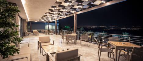 Een terras buiten met zitplaatsen en tafels, een uitzicht op de stad 's nachts, en een modern plafondontwerp.