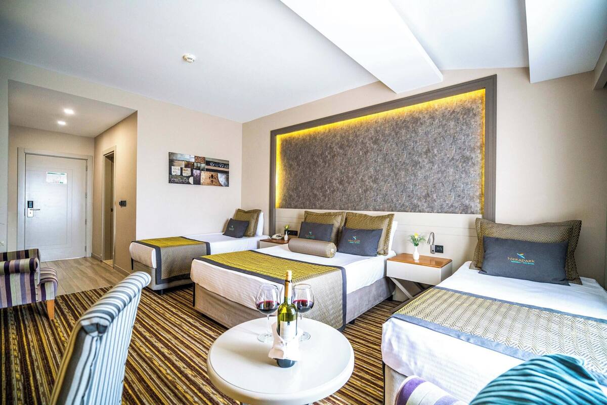 Habitación de hotel con una cama, dos mesitas de noche, un sofá, una mesa de centro con copas de vino y una obra de arte colgada en la pared.