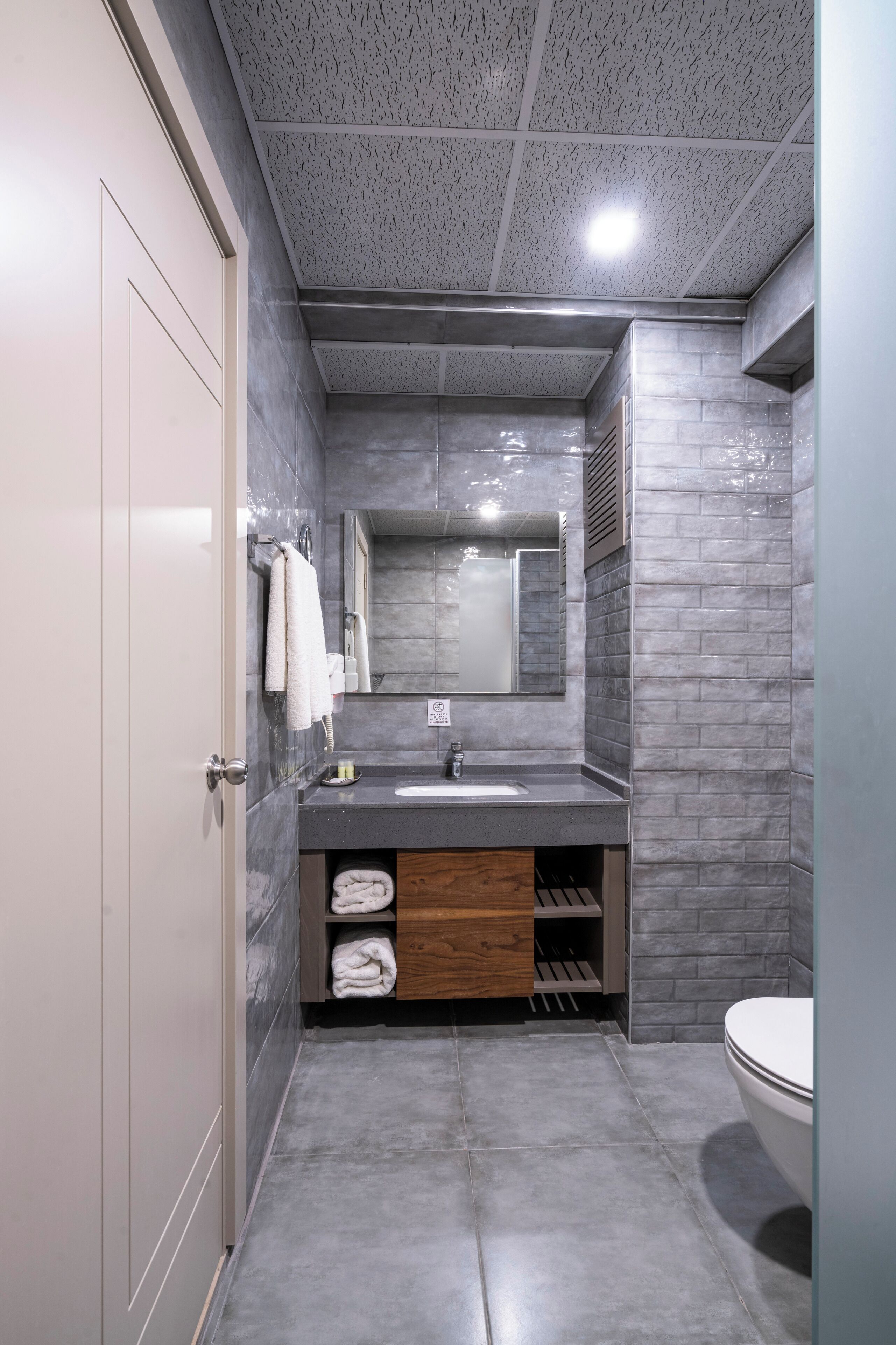 A modern bathroom with a grey tiled floor, a white toilet, a grey sink with a mirror above it, and a towel rack with white towels.