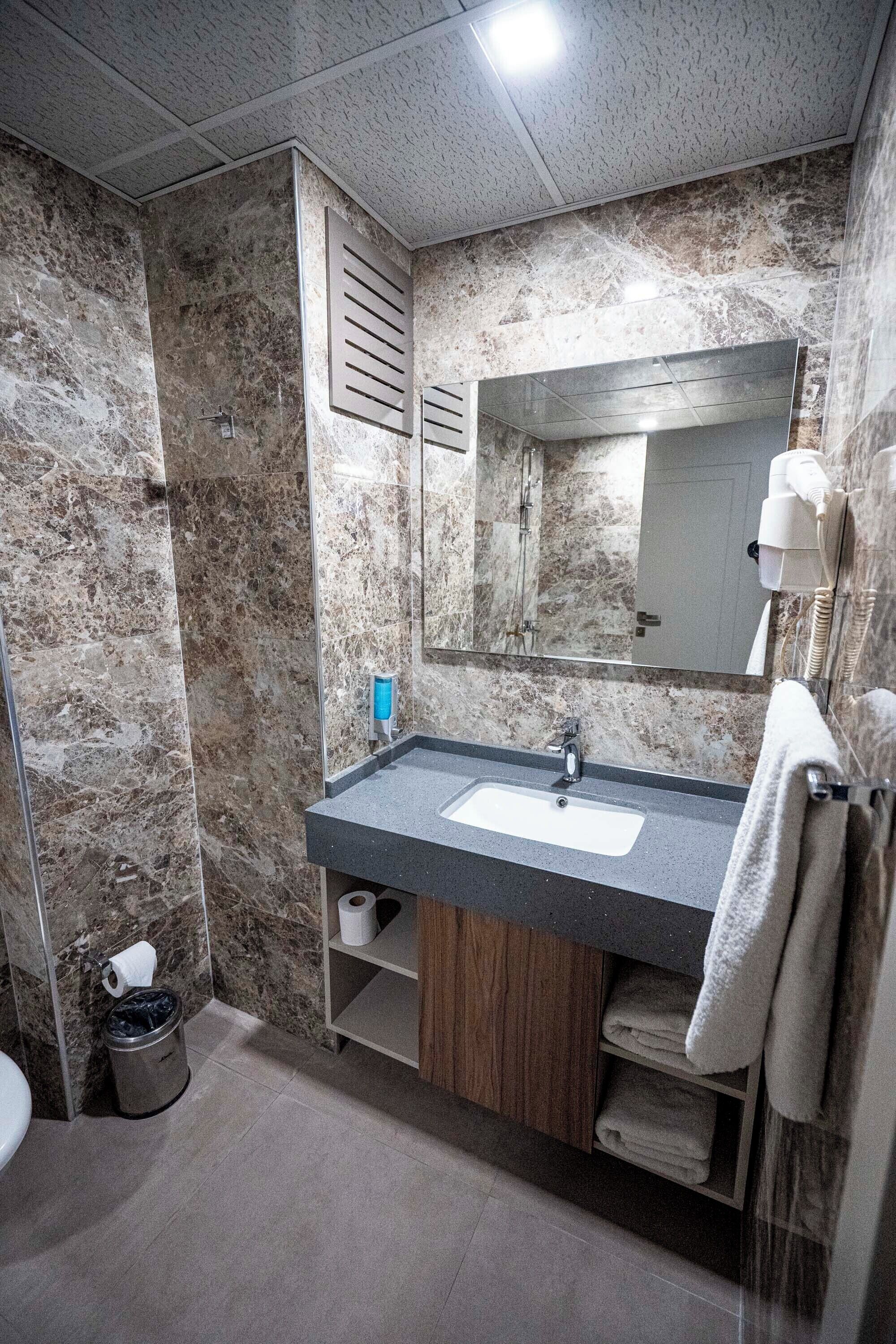 Un baño con pared de mármol, un lavabo con espejo, un perchero y un cesto de basura.