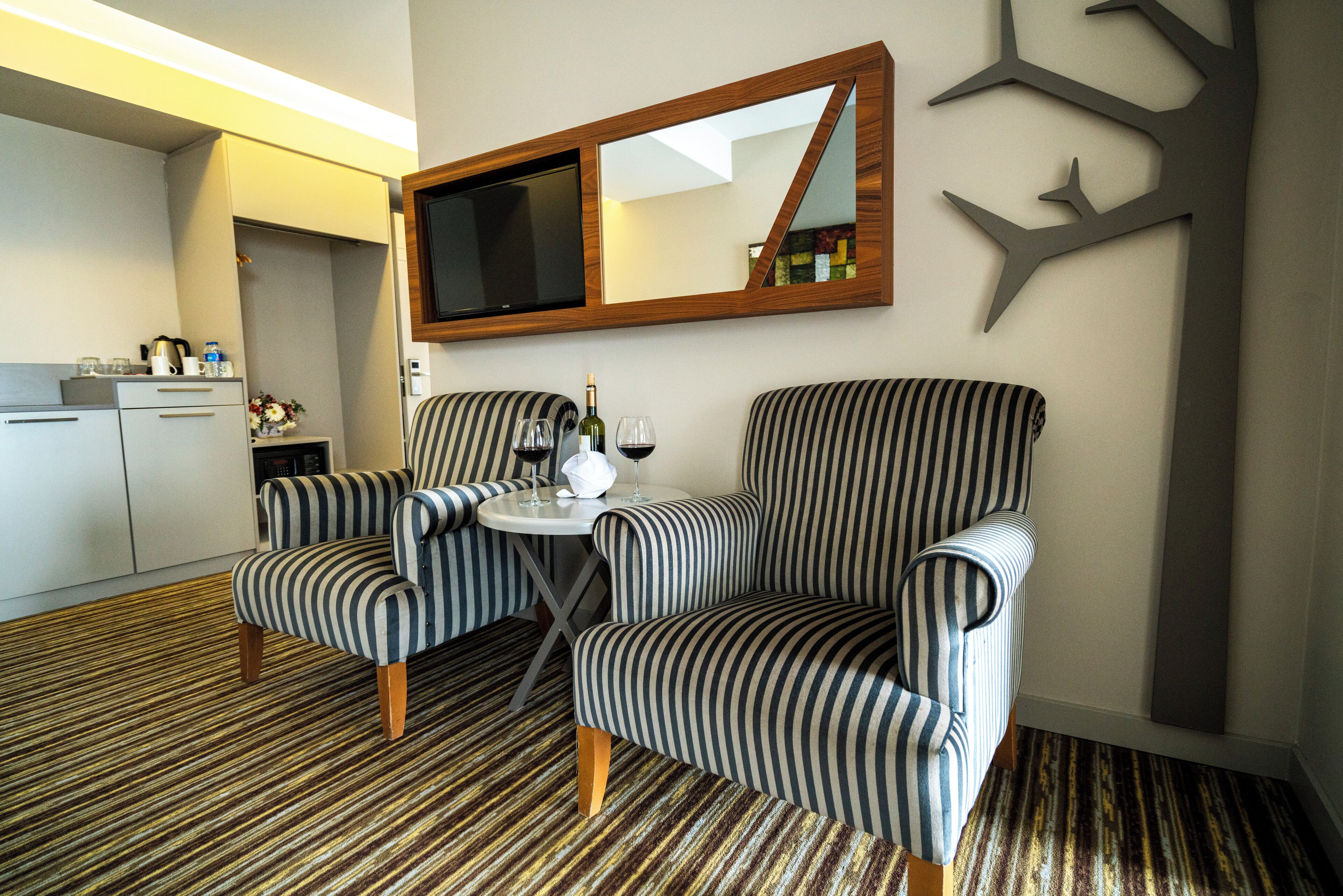 A hotel room with striped armchairs, a small table with a bottle and glasses, and a TV mounted on the wall.