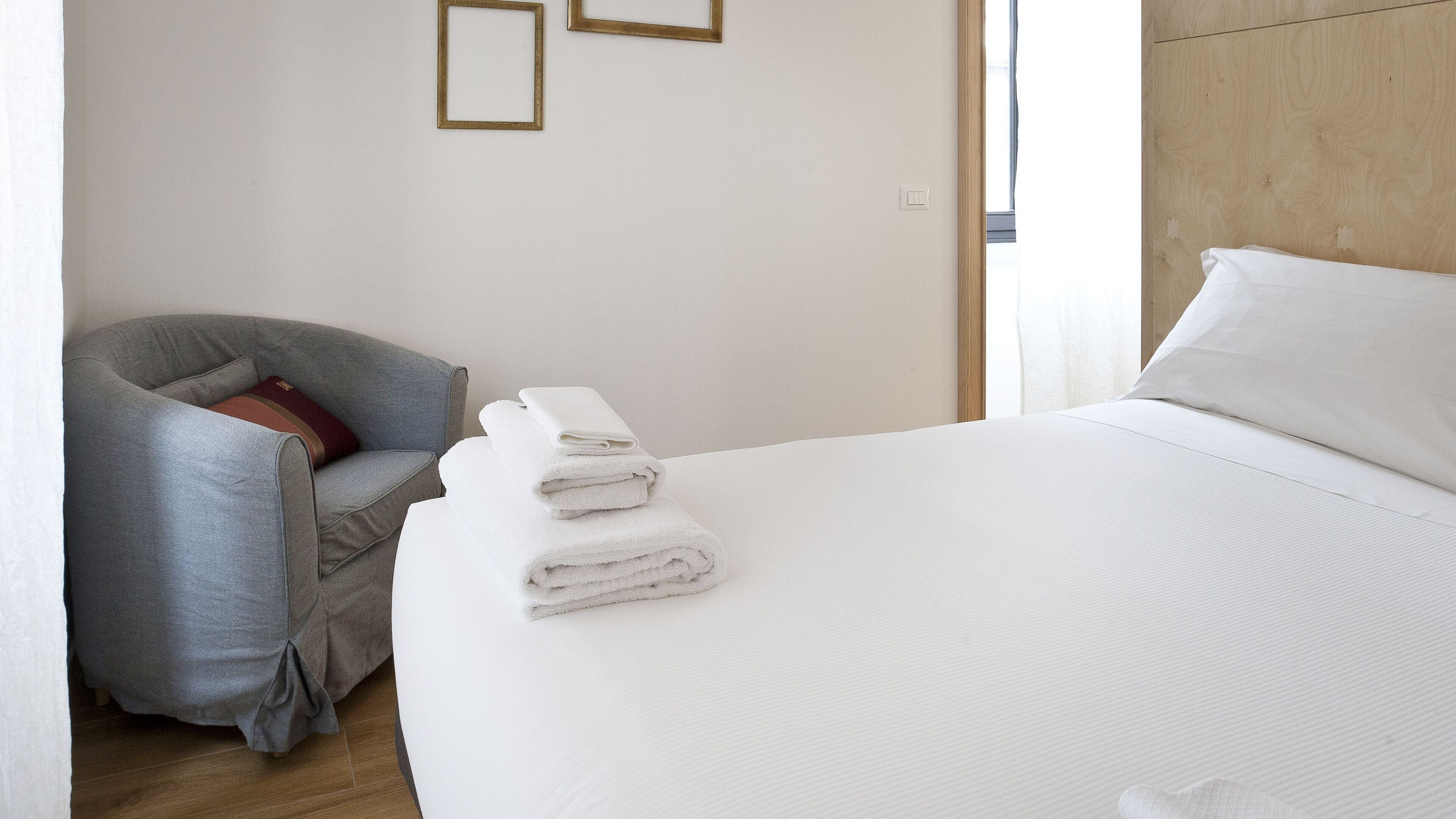 A hotel room with a bed, a grey armchair, and a stack of white towels.