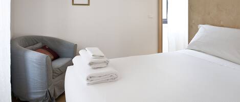 A hotel room with a bed, a grey armchair, and a stack of white towels.
