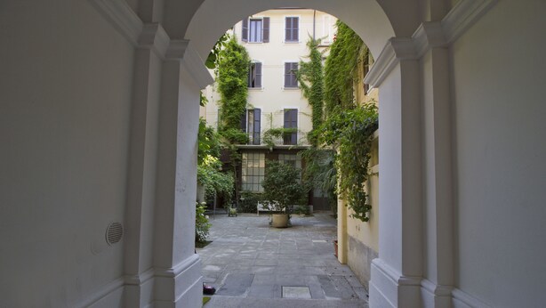 A narrow courtyard with a central planter, flanked by buildings with windows and vines. - Italianway   - Caretto (Milan)