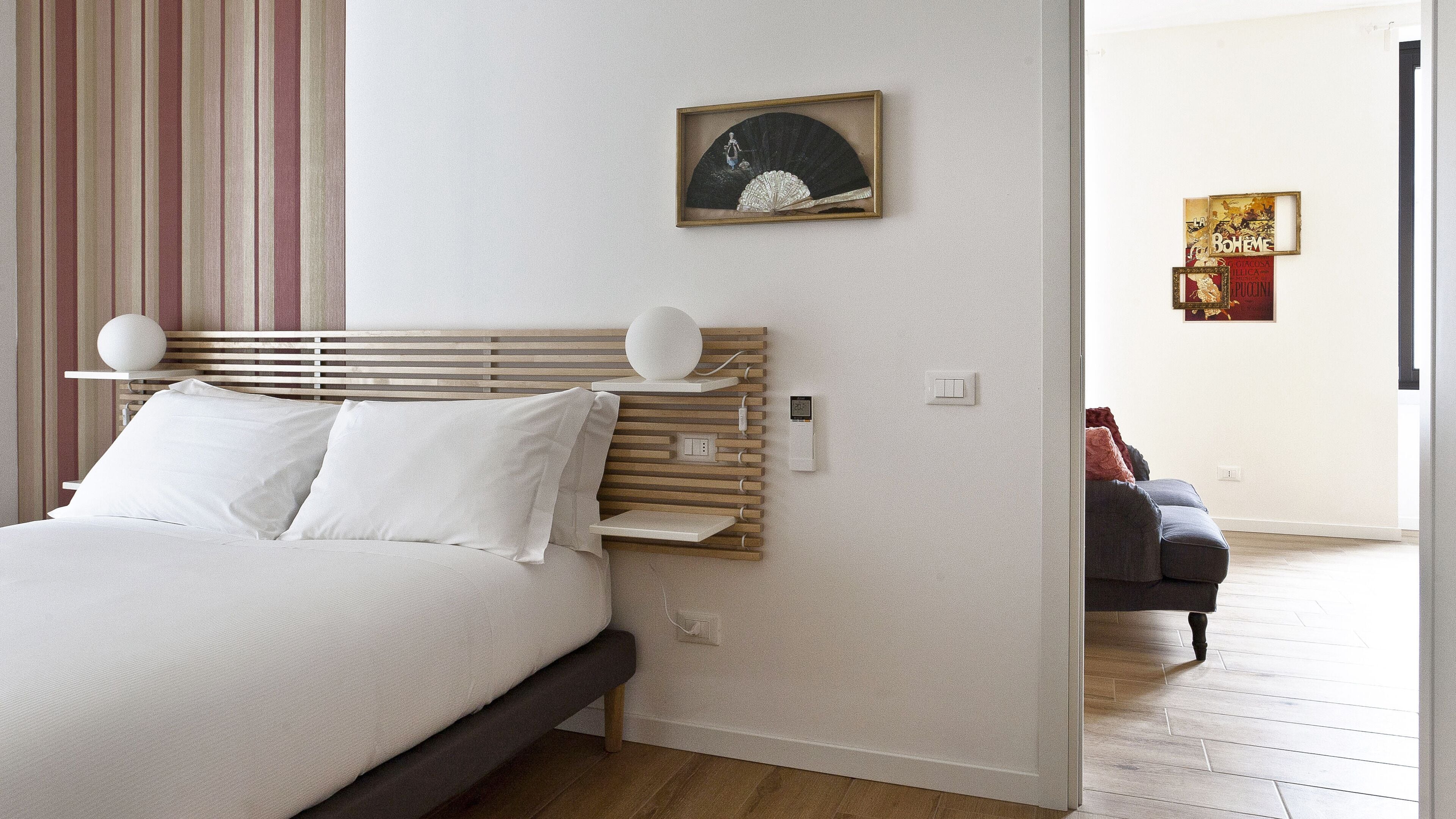A modern bedroom with a wooden headboard, a bed with white bedding, a bedside table, and a wall-mounted light fixture.