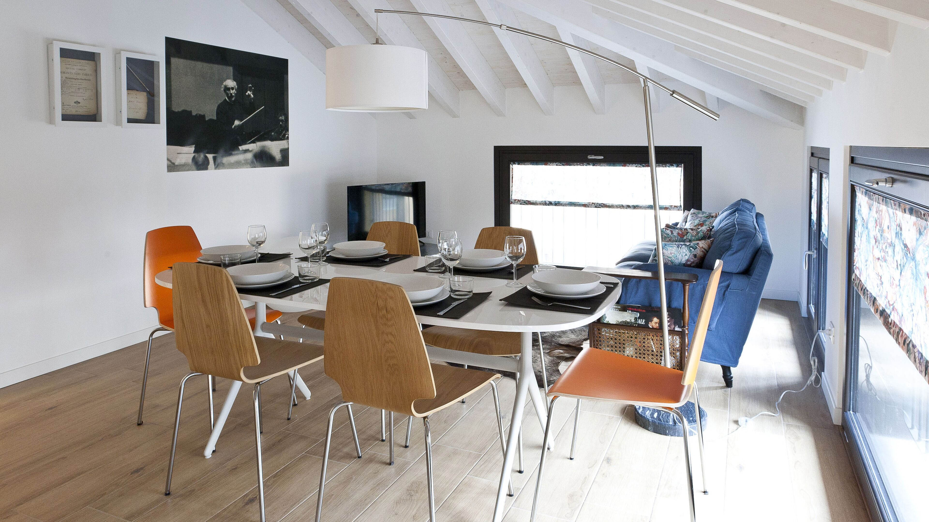 Un comedor con una mesa blanca dispuesta para cuatro personas, rodeada de sillas de madera. Un televisor está montado en la pared, y hay una ventana que proporciona luz natural.